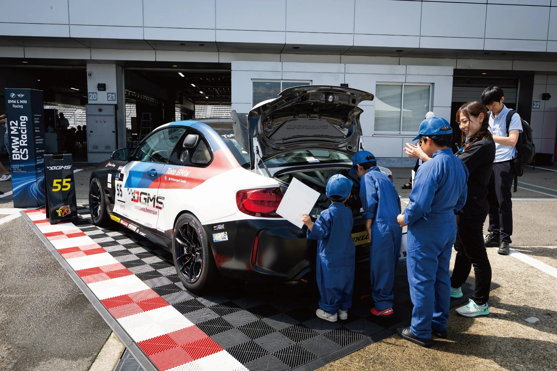 サーキットのピット前で、BMW M2 CS Racingカーのトランクを覗き込む子供たち
